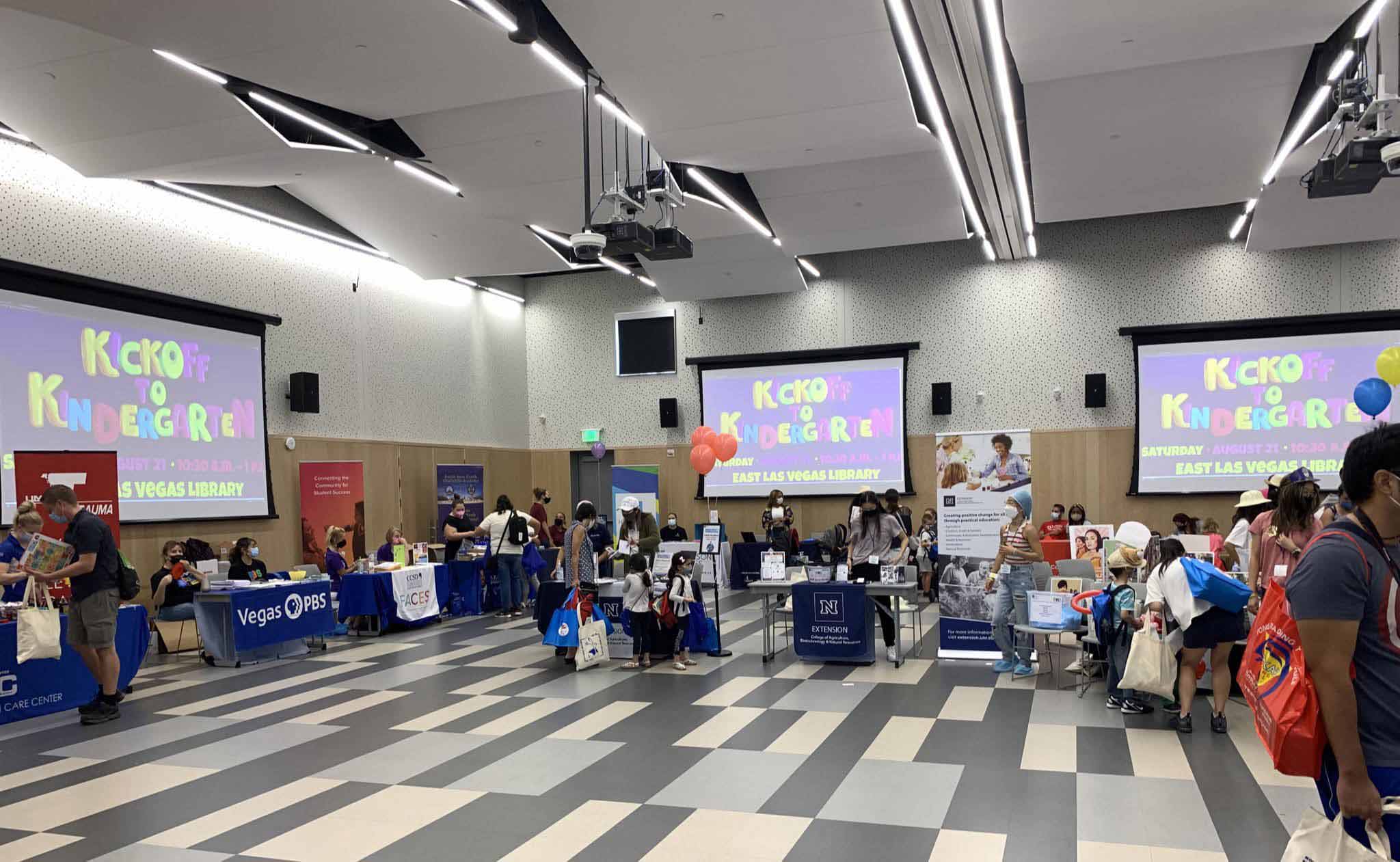 A large, open room is flanked with colorful tables and booths staffed by masked community partners. Masked families and children are exploring the booth and participating in the activities. On several projector screens in the room are the words Kickoff to Kindergarten in a colorful, child-like font.