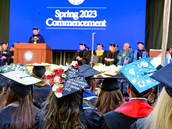 In the distance, University President Brian Sandoval is addressing graduates from the 2023 Spring Commencement stage. In the foreground are students in academic regalia. Two decorated caps read: Para mis padres que llegaron sin nada y me lo dieron todo" and "I already forgot everything."