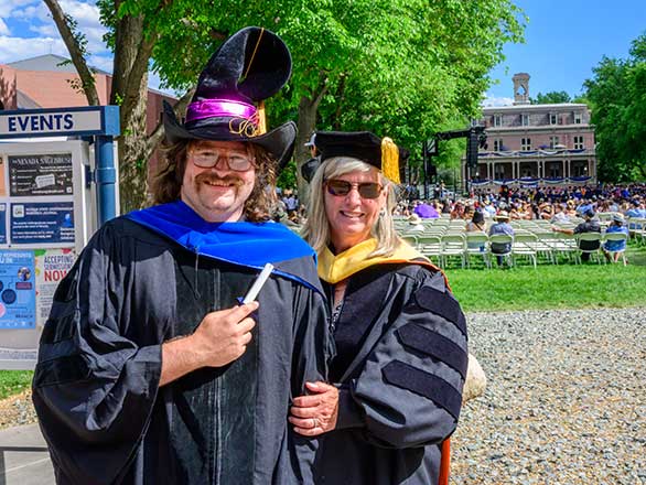A professor and student in academic regalia on the University quad. The student is holding a scroll and wearing a tall black had that looks like a cross between a magician's hat and a cowboy hat.