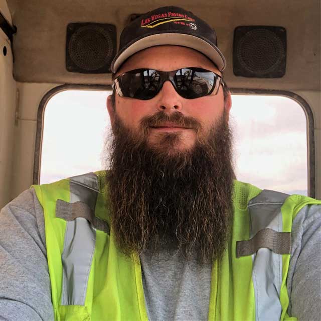 Dusty Domina in sunglasses, cap and hi-viz vest, sitting in the cab of a piece of heavy equipment