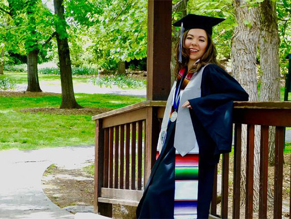 Ana in graduation regalia leaning against a wooden railing, green trees in the background