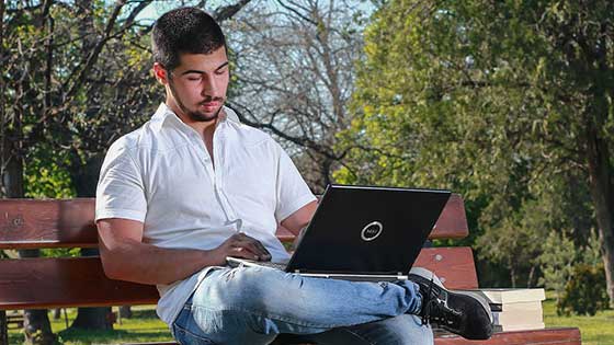 Student using a laptop on a bench, blue skies, tall trees and green grass in the background
