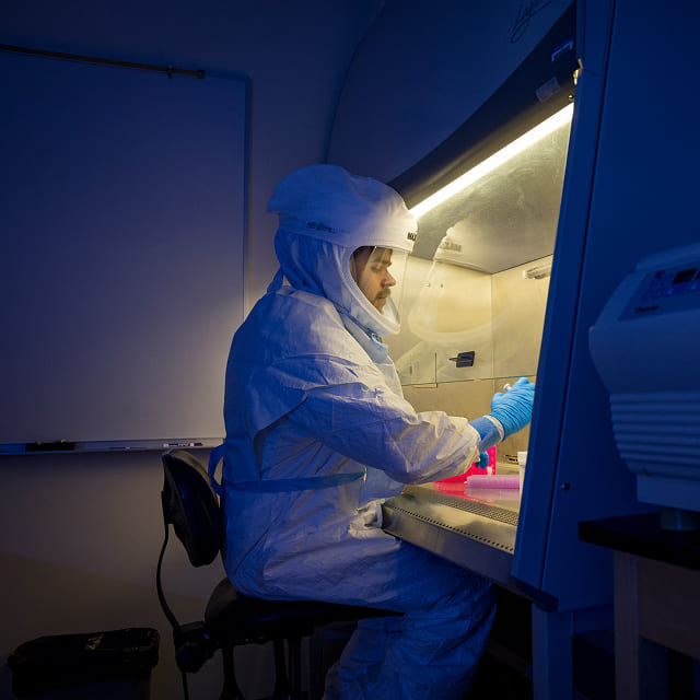 A researcher looks at a sample while wearing full protective gear