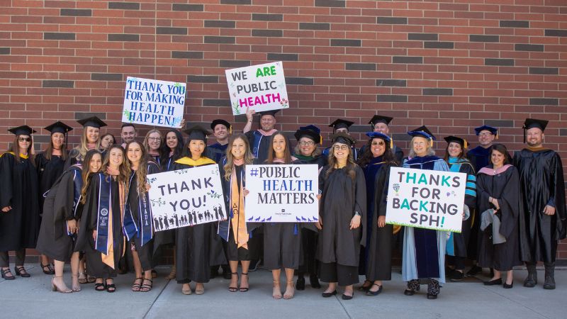 A group of students and faculty at commencement, dressed in their cap and gowns while holding public health signs.