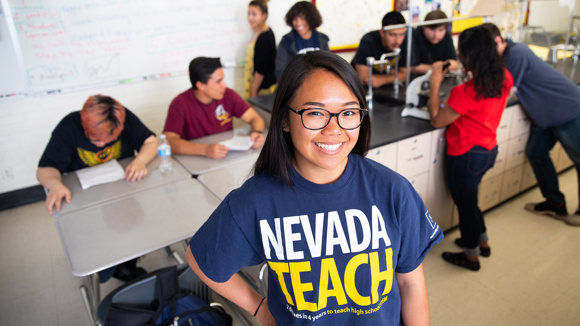 A person wears a NevadaTeach shirt while standing in front of other people seated at a desk.