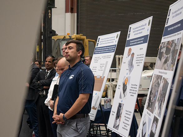 A man wearing stands in front of posters about the Laminar Soil Box mounted on easels, looking at something out of the photo frame, to the left, with five people in the backround, also looking left.