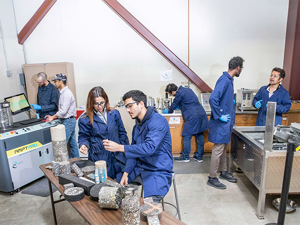 Two researchers in blue lab coats look as asphalt mix samples while five people in the backgroun work at machines.