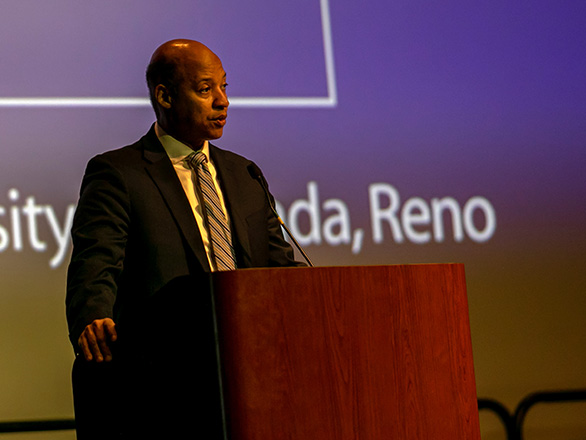 Erick Jones in suit standing at a podium, screen in the background on which part of the phrase "University of Nevada, Reno" is visible.