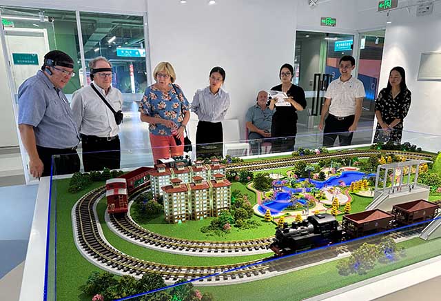 Eight people looking at a model train; the two people on the left are wearing headsets that control the movements of the train.