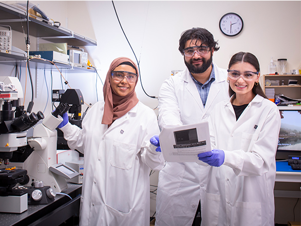 Three students in lab coats and goggles post in a lab next to microscopes.