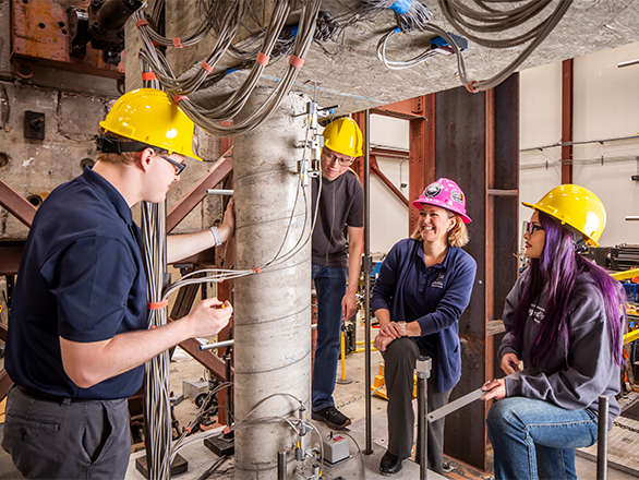 A professor and three students in hard hats gather around a concrete pillar being tested on an earthquake shake table.