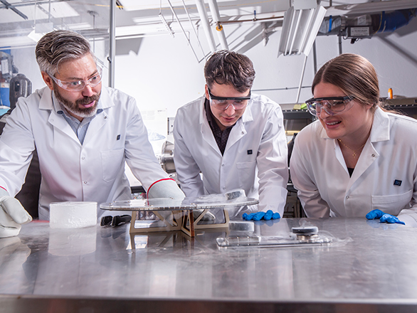 A faculty member and two students wearing lab coats and protective examine components and materials on a stainless steel workbench.