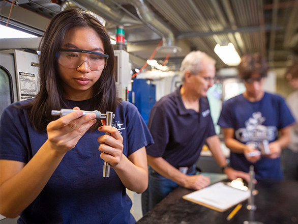 A female student wearing protective goggles works with two metal tubes while a faculty member and other students talk in the background