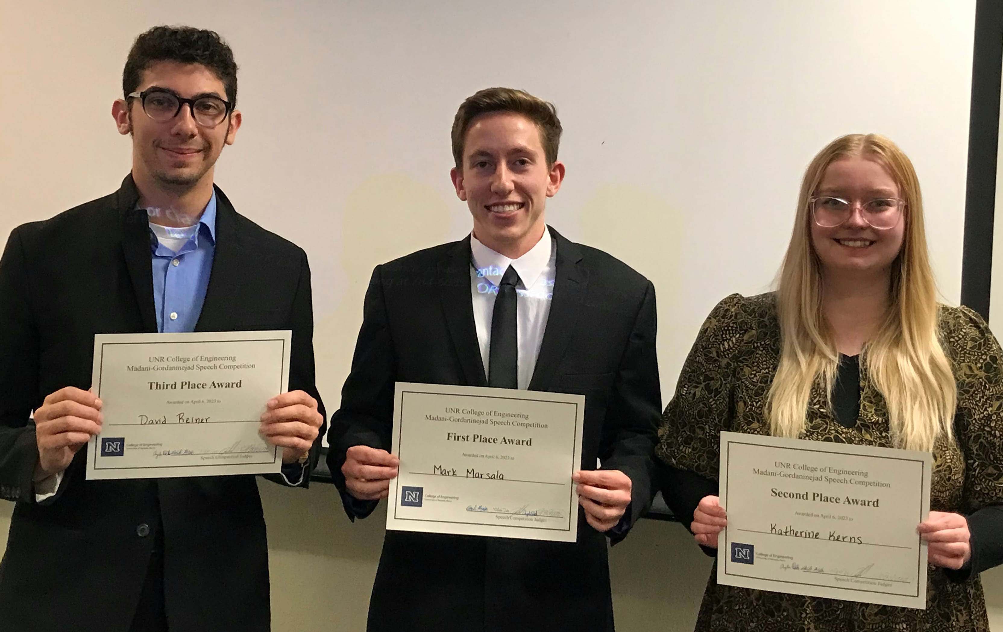 Three people standing in a row, indoors in front of a white wall, each facing the camera and holding a certificate.