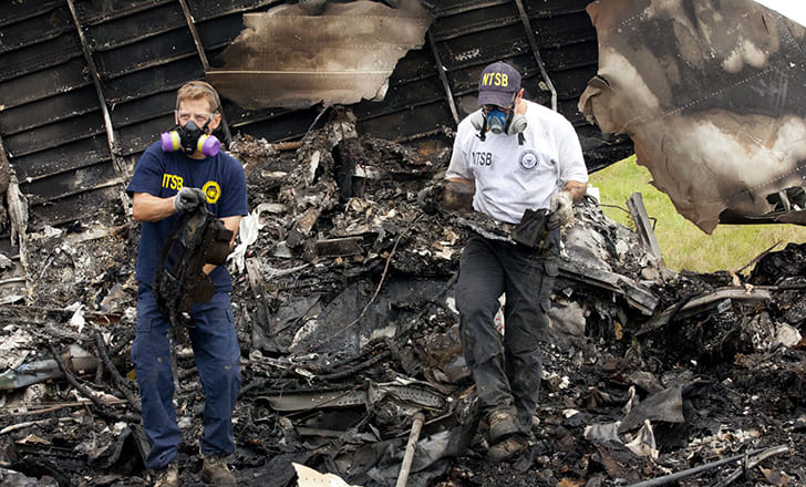 Two people standing on burned crash debris, holding hunks of metal and wearing face masks