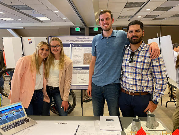 Four students, two female, two male standing in front of a project poster, with a table in front of them.