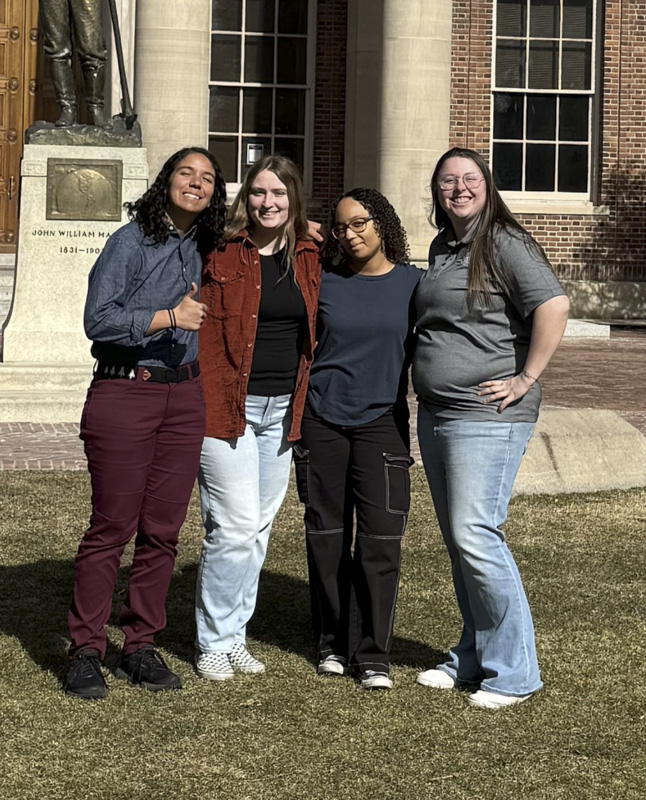 Four women stand in front of a brick building with columns, smiling at the camera.