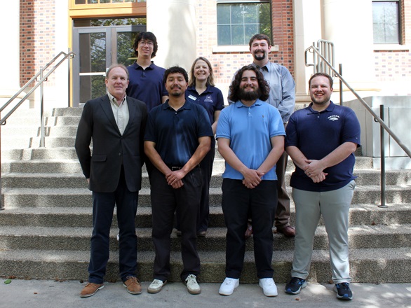Six people stand in two rows, four in front, two in back, on a cement staircase in front of Palmer Engineering Building