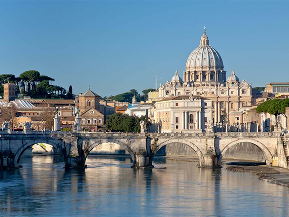 Ornate historical building with a domed roof bordered by other buildings sits on the edge of a river spanned by a bridge.