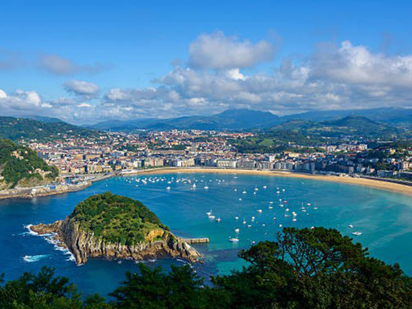 View of a bay filled with sailboats, and island in the foreground and beach and city in the background.
