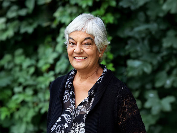 Woman wearing a black-and-white blouse and a white sweater smiling, greenery in the background.