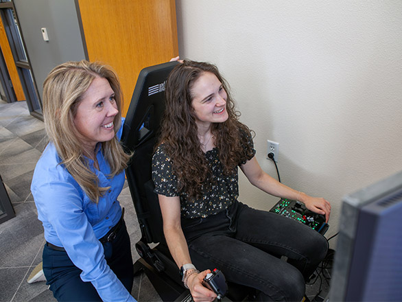 A woman sits in a flight simulator, with another woman kneeling behind her.