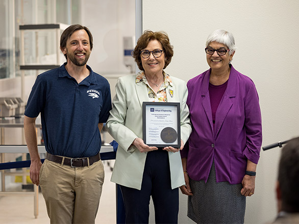 Three people, one holding a certificate, state in front of windows looking into a laboratory.
