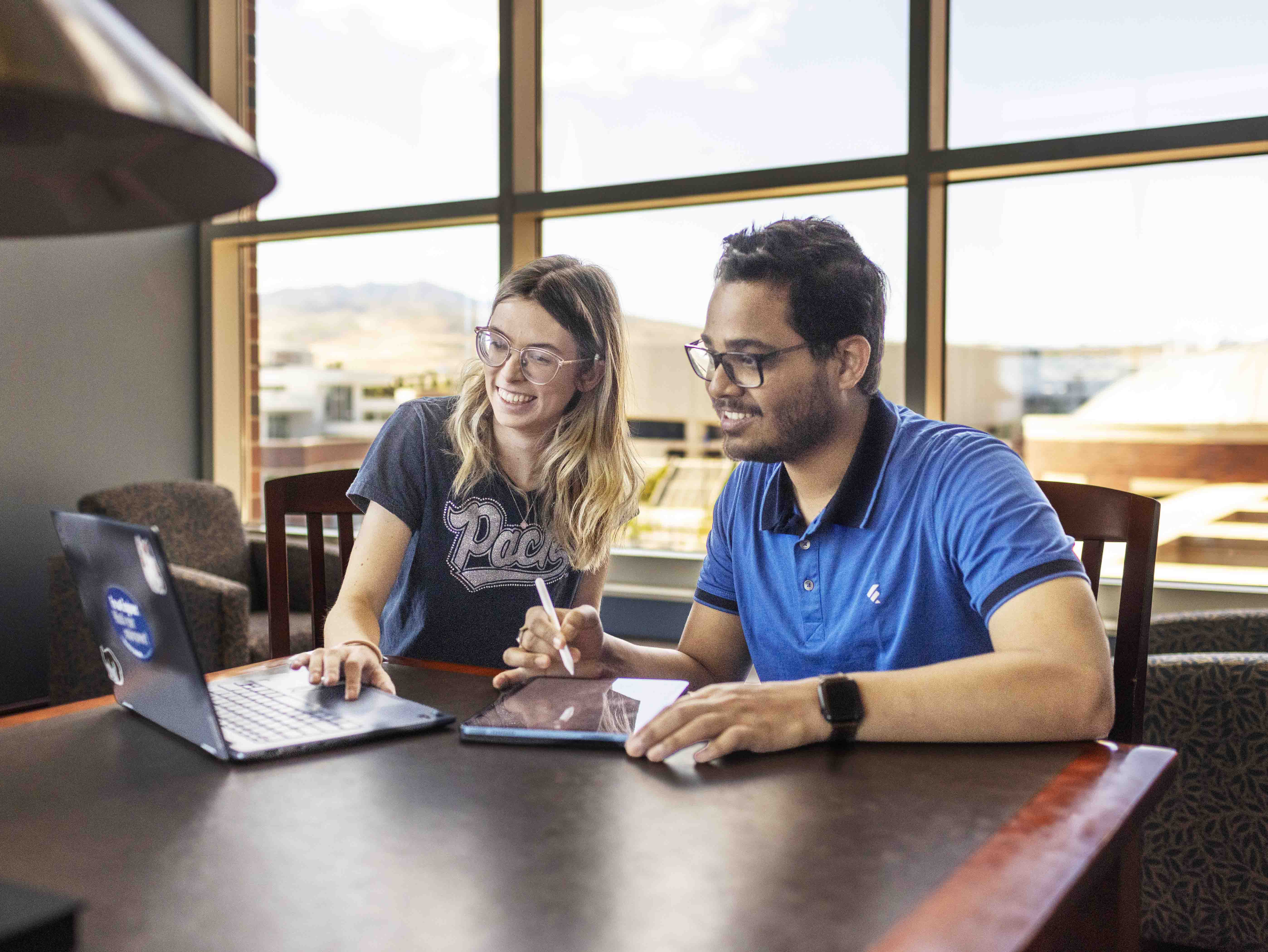 Two students sit at a library table, looking at a laptop.