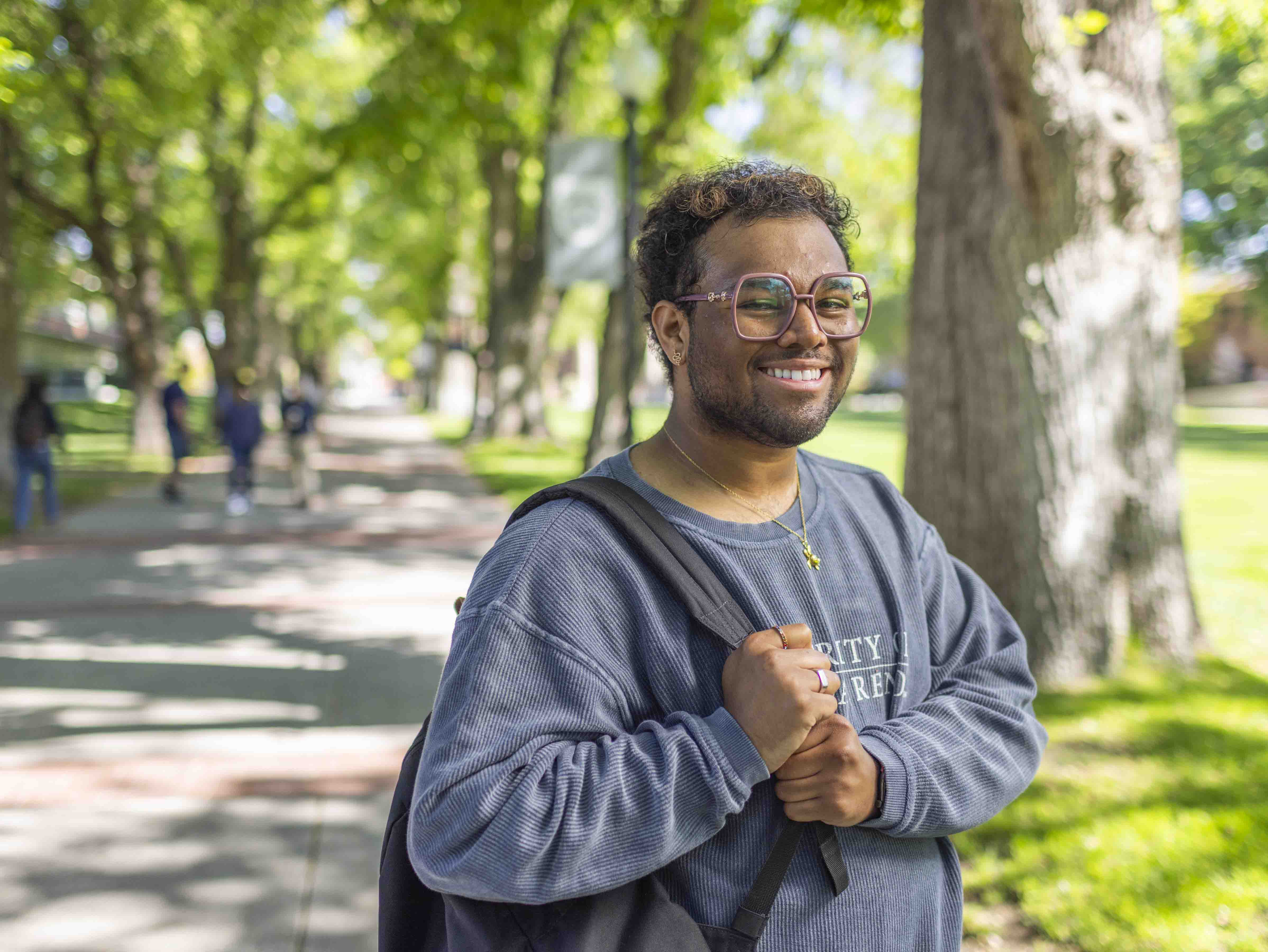 Male student wearing a University of Nevada, Reno sweatshirt and holding a backpack stands in front of trees and a walkway, with people in the background.