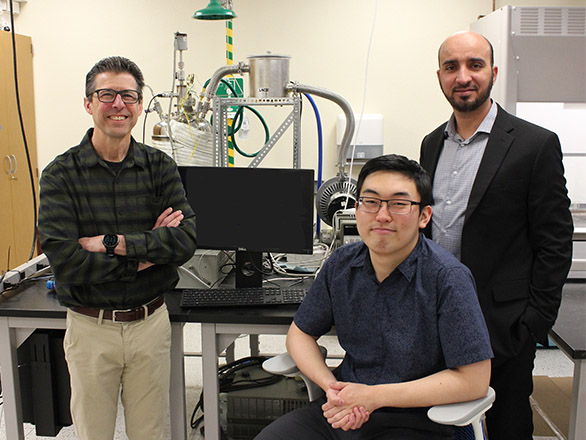 Two faculty members, smiling at the camera, stand behind a seated student in a lab.