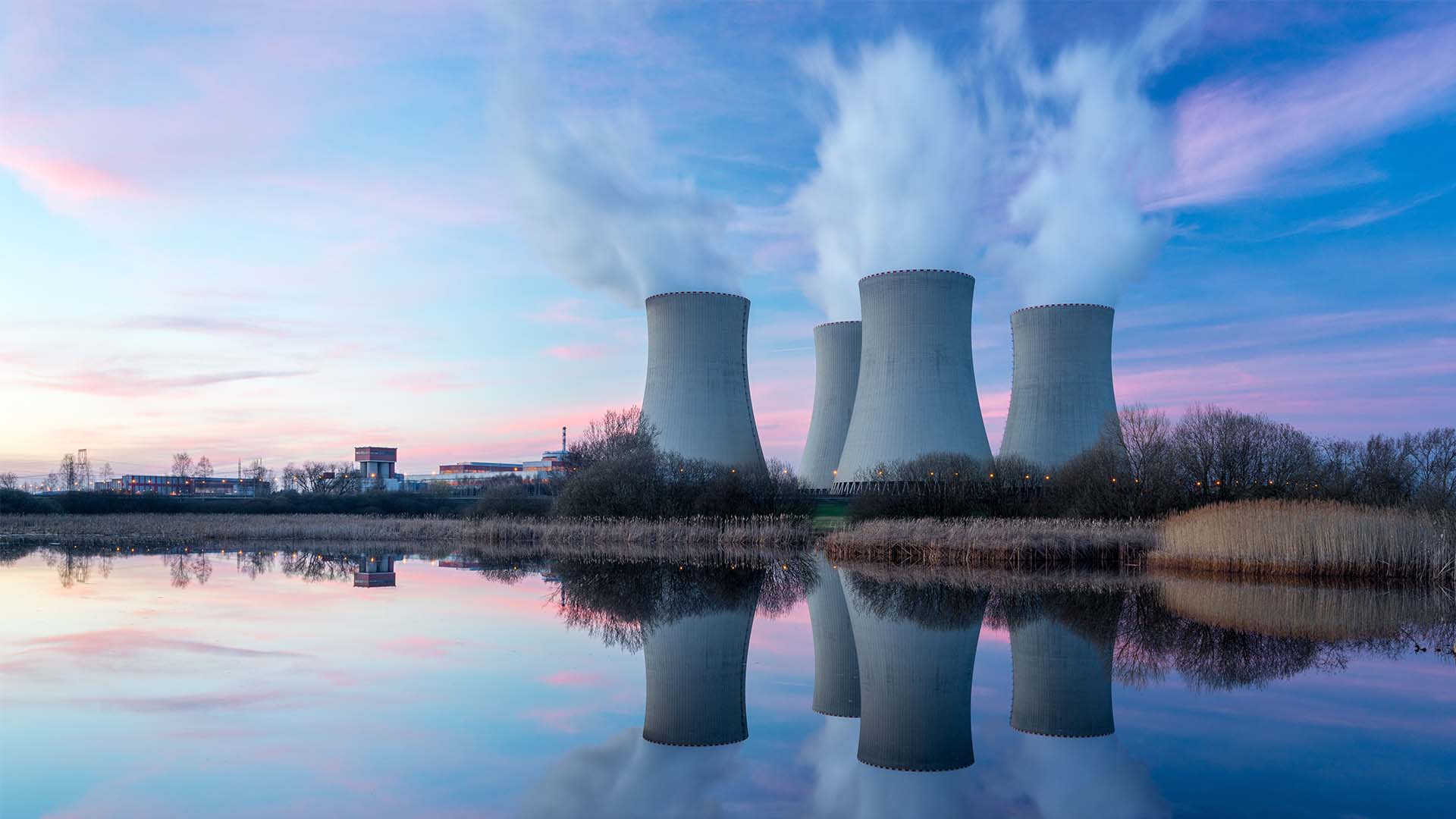 Landscape of a power plant with steam or smoke rising from four columns and a body of water in the foreground.
