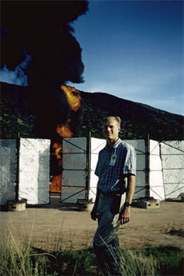 Person at test site with large fire and partition behind