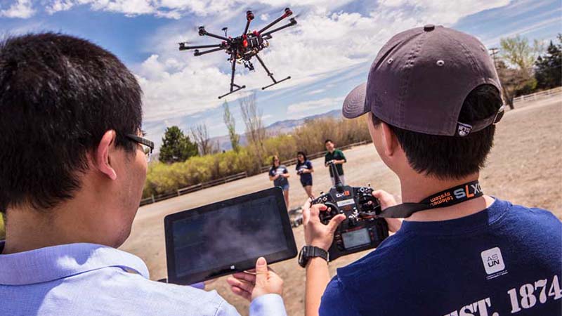 Two students facing away from camera operating an aerial drone that hovers between them.