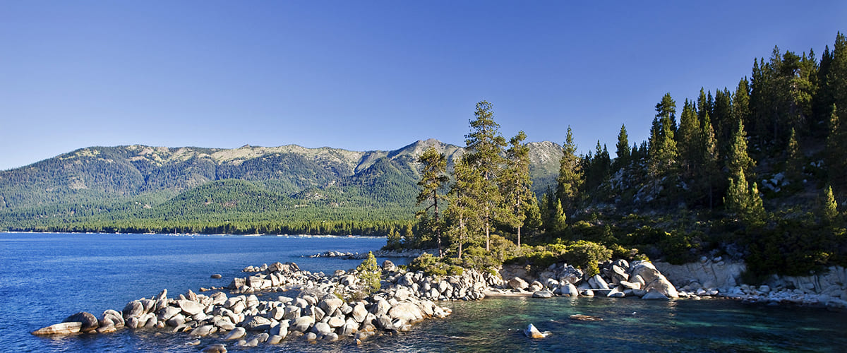 A view of Lake Tahoe, with forested mountains in the background and rocks spilling out in the lake 