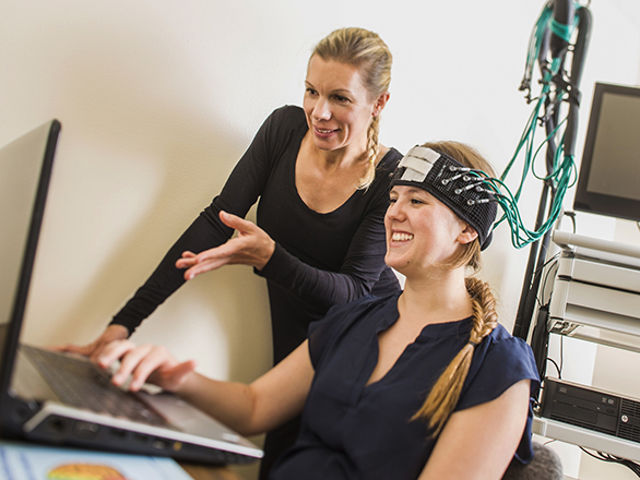 Two researchers are reviewing content on a computer while one is wearing monitoring sensors on their head. 