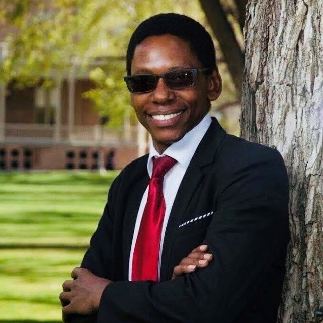 Liberatus J. Rwebugisa wearing sunglasses and leaning against a tree