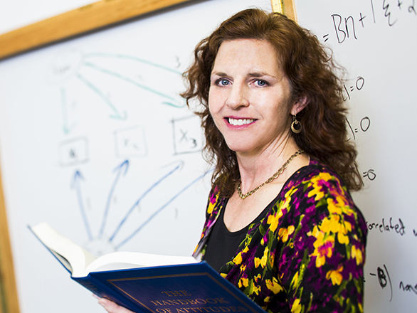 A faculty member smiles and holds a book while standing in front of a white board.