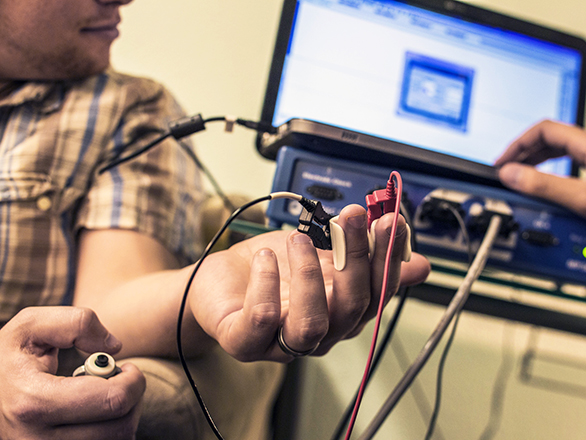 A person sits with an electronic measuring device attached to their fingertips while another person monitors results on a computer. 