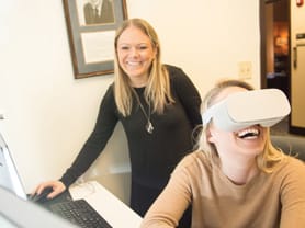 A faculty member clicks a mouse controlling a headset worn by a student in the CAMS lab.