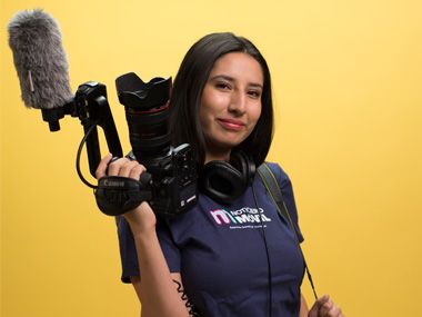Student holds video camera and wears a Noticiero Móvil shirt.