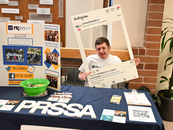 A student tabling at a club fair while holding a framed poster of an social media post. 