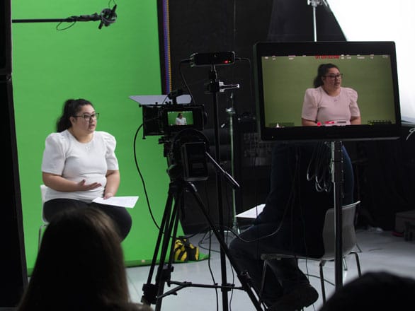 A student speaks to a professor while sitting in front of a green screen and being recorded on video.
