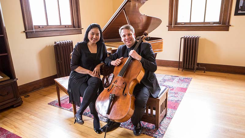 Dmitri Atapine poses with a cello sitting next to Hyeyeon Park on a piano bench, with a piano behind them.