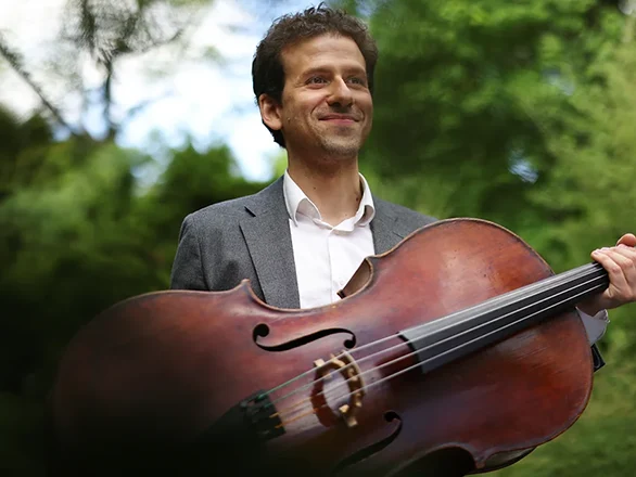 Nicholas Canellakis wears a gray suit and holds a cello sideways while posing for a picture in front of some trees.