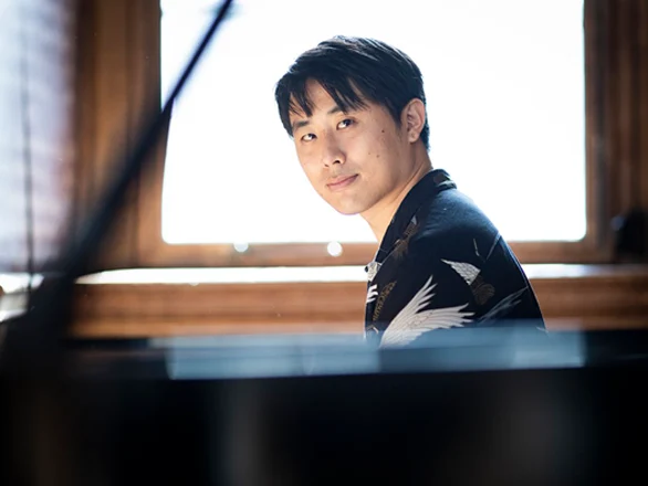 George Fu wears a shirt with images of birds while sitting at a piano with a window behind him.