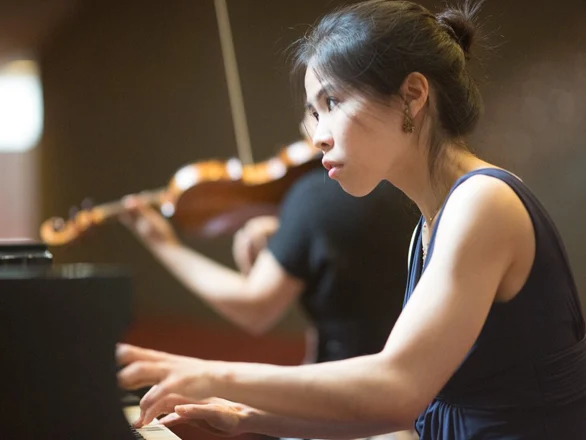 Mika Sasaki plays the piano while a person plays a violin behind her.