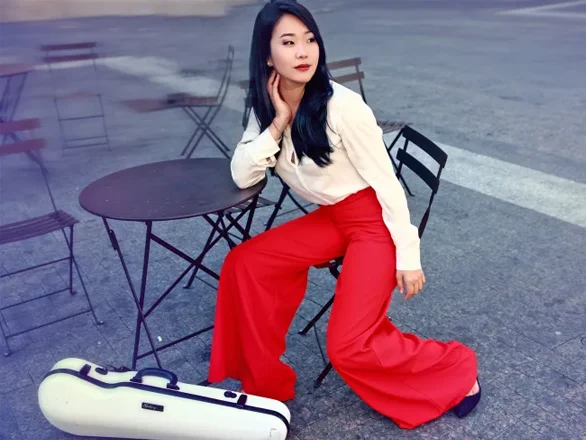 Kristin Lee wears a white blouse and red pants and sits on a chair at a table with a violin case at her feet.