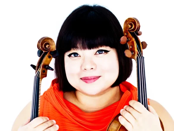 Yura Lee wears a red shirt and poses for a photo while holding the handles of a violin and viola in each hand.