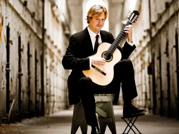 Jason Vieaux wears a suit and sits on a stool playing a guitar in an older building.