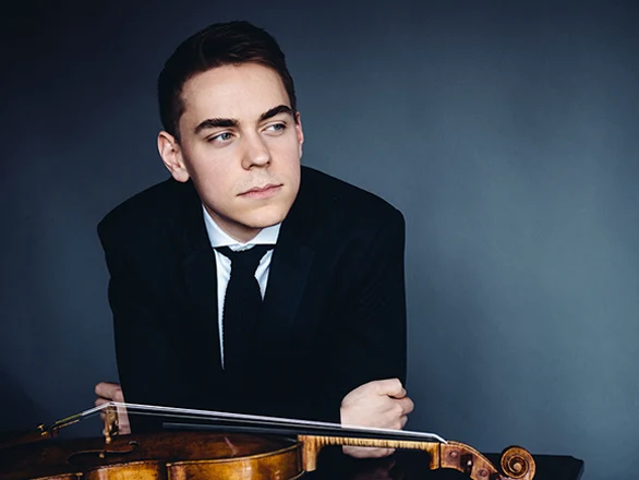 Matthew Lipman wears a suit and rests with his arms folded on a table with a viola resting in front of his arms.